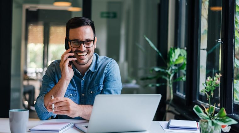 Portrait of young smiling cheerful entrepreneur in casual office making phone call while working with laptop