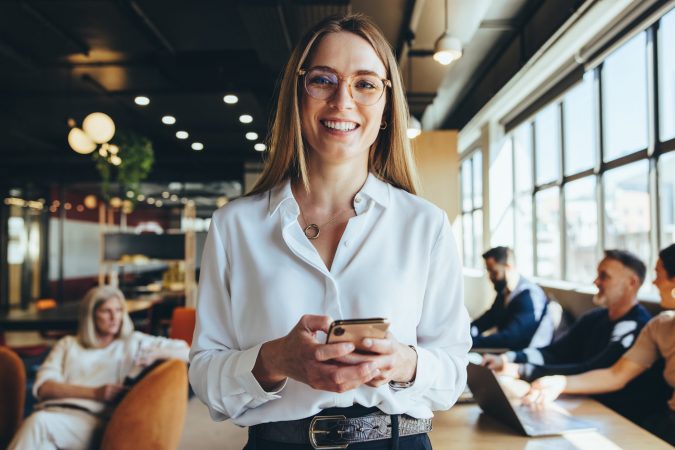 Young businesswoman holding a smartphone in a co-working space. Happy young businesswoman smiling at the camera while standing in a modern workplace. Female entrepreneur sending a text message.